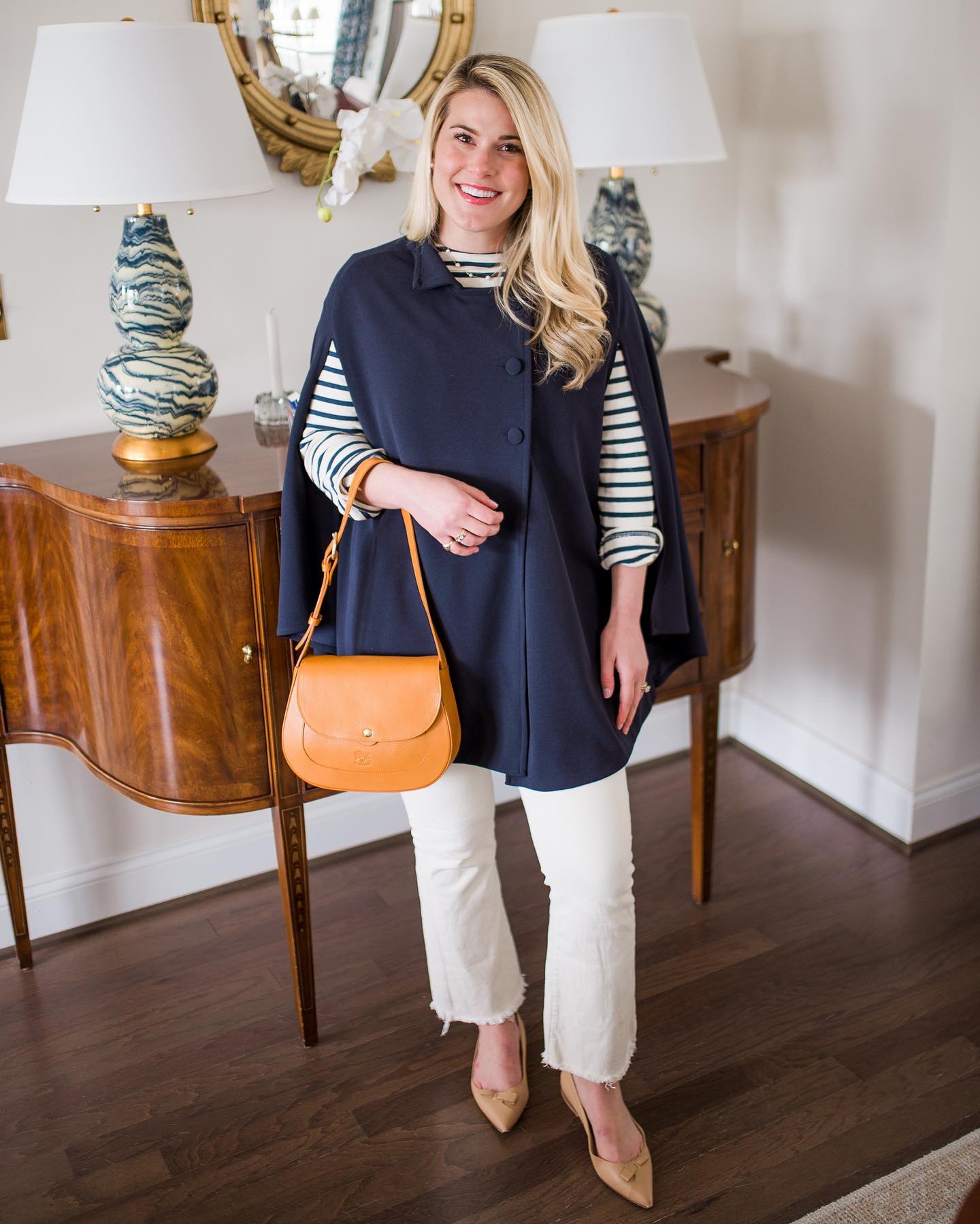 Smiling blond woman wearing Marta Scarampi's navy blue cape. The woman looks into the camera and lifts her leg slightly, showing her comfortable heels and walking bag. 
