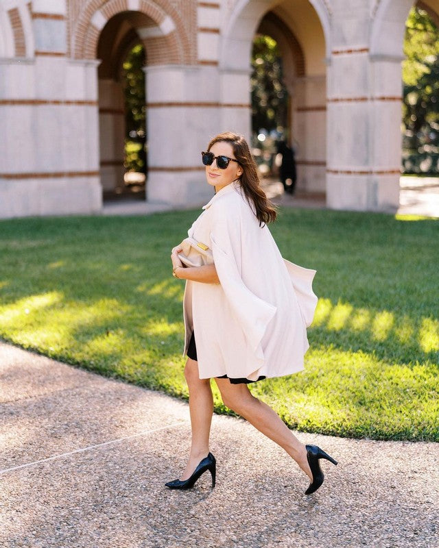 Elegant brunette woman wearing Rachel Mondrian Cape in cream colour. The woman is looking into the camera, wearing sunglasses and elegant black heels. The pose is in motion so the cape gives the woman wings. 
